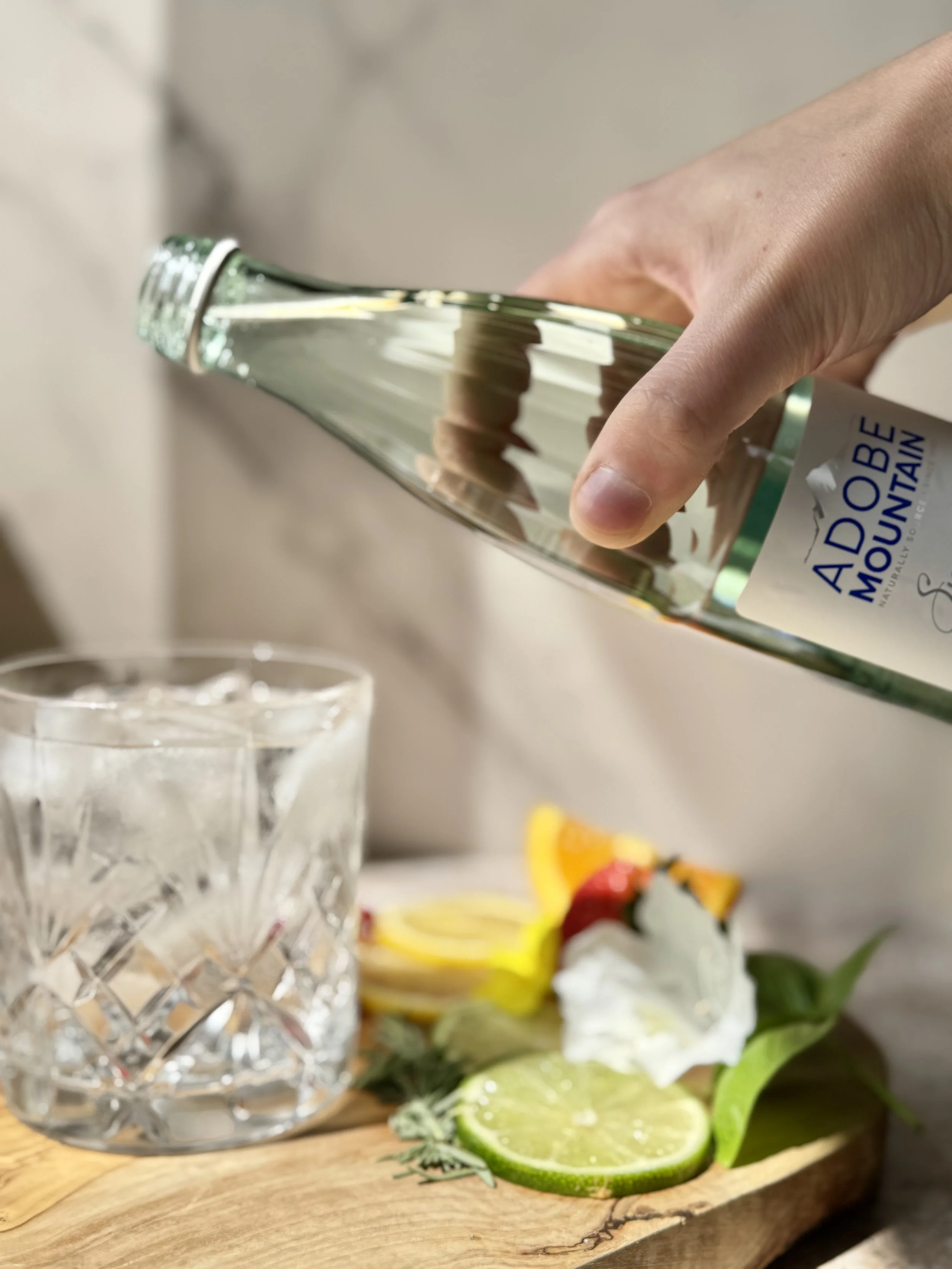 A hand tipping an Adobe Mountain bottle toward a cut-glass tumbler beside sliced citrus and herbs