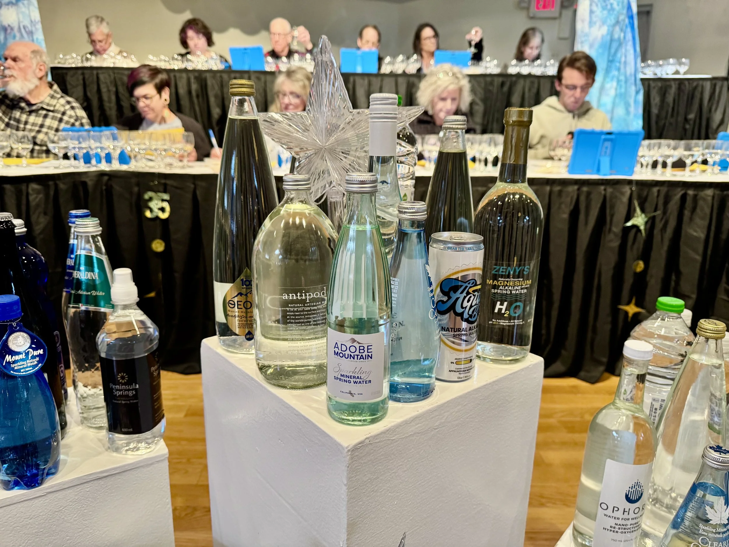 An Adobe Mountain sparkling bottle on a display pedestal among other waters, with a glass star and tasting judges behind it